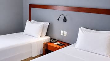 A hotel room with two single beds, a wooden headboard, a bedside table with a telephone and remote controls, and a wall-mounted light.