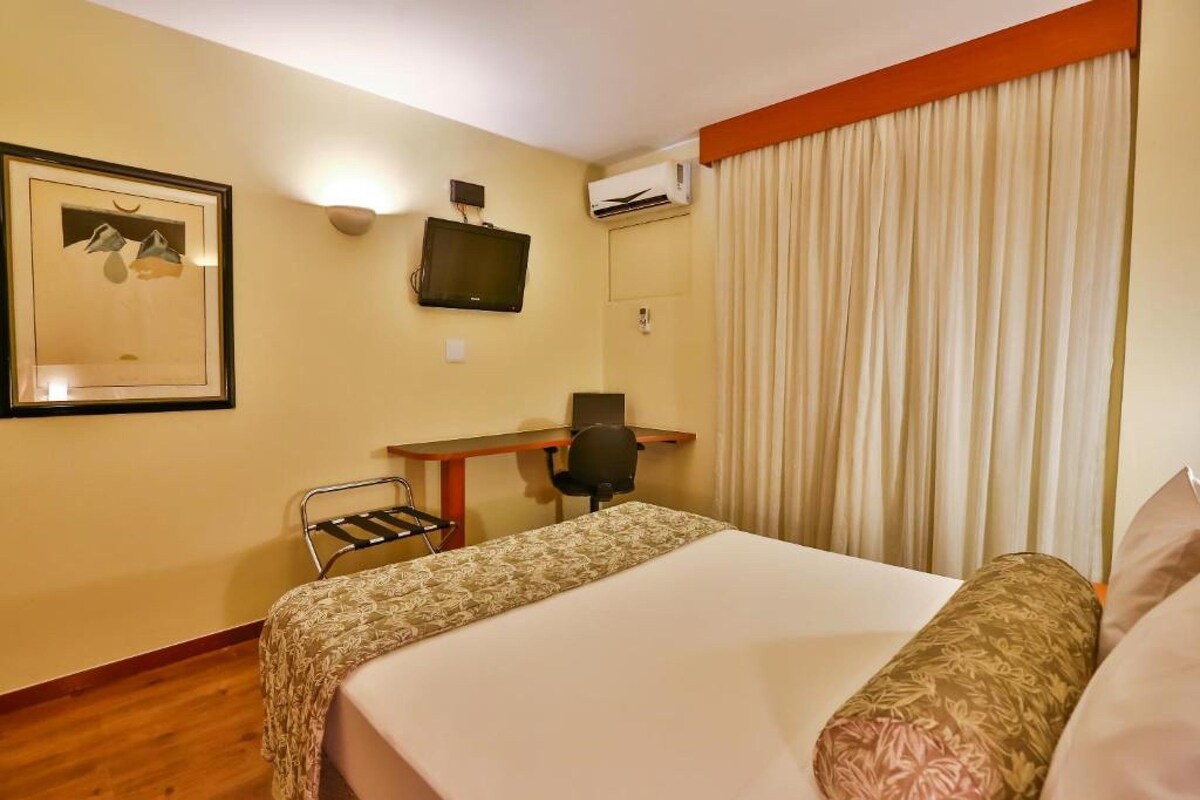 Habitación de hotel con cama, escritorio, silla, unidad de aire acondicionado, televisor y un cuadro en la pared.