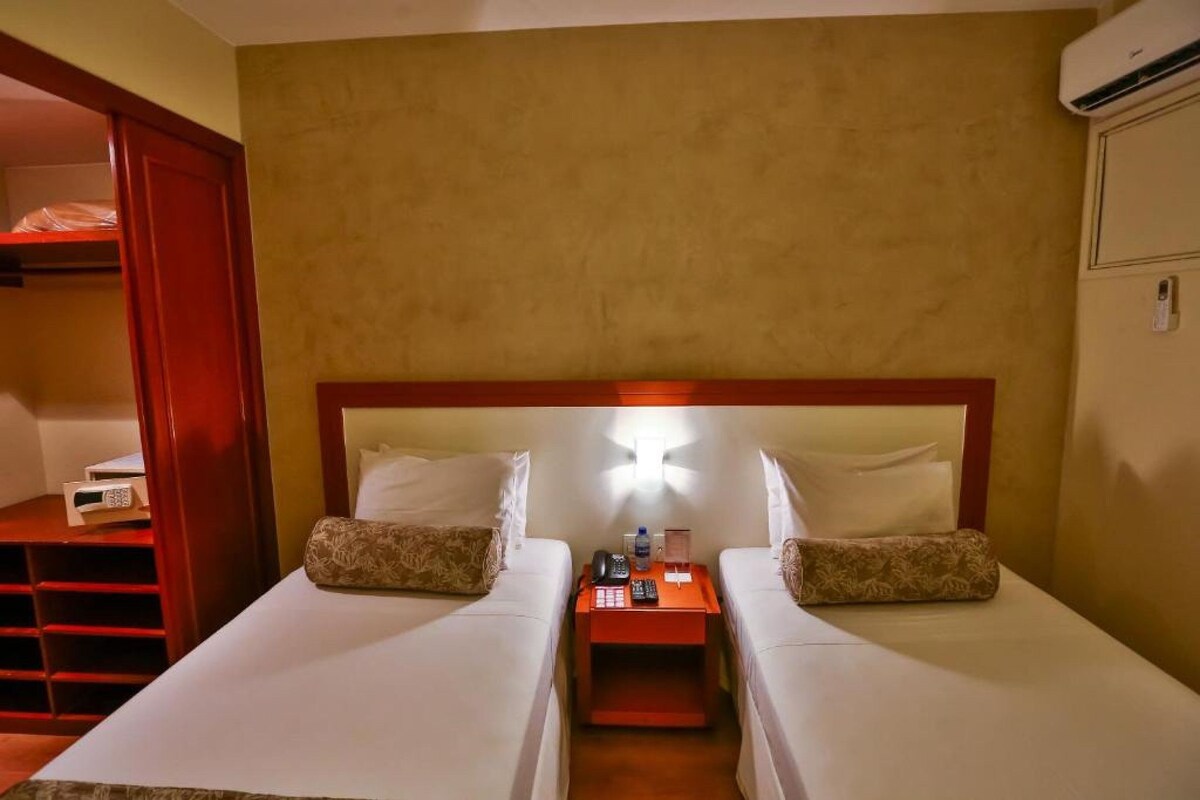 Habitación de hotel con dos camas individuales, una mesita de noche con lámpara, una mesita auxiliar roja y un equipo de aire acondicionado en la pared.
