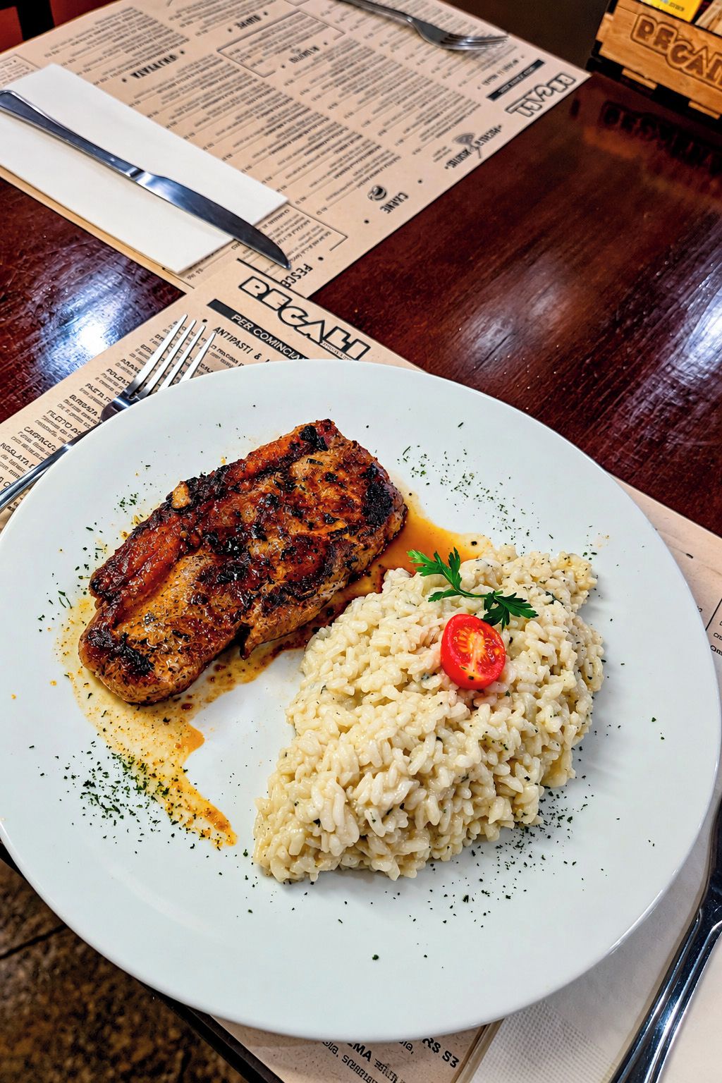Prato com carne grelhada e risoto, um menu e talheres sobre uma mesa de madeira.