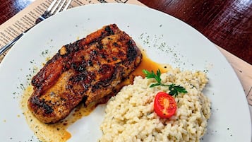 Prato com carne grelhada e risoto, um menu e talheres sobre uma mesa de madeira.