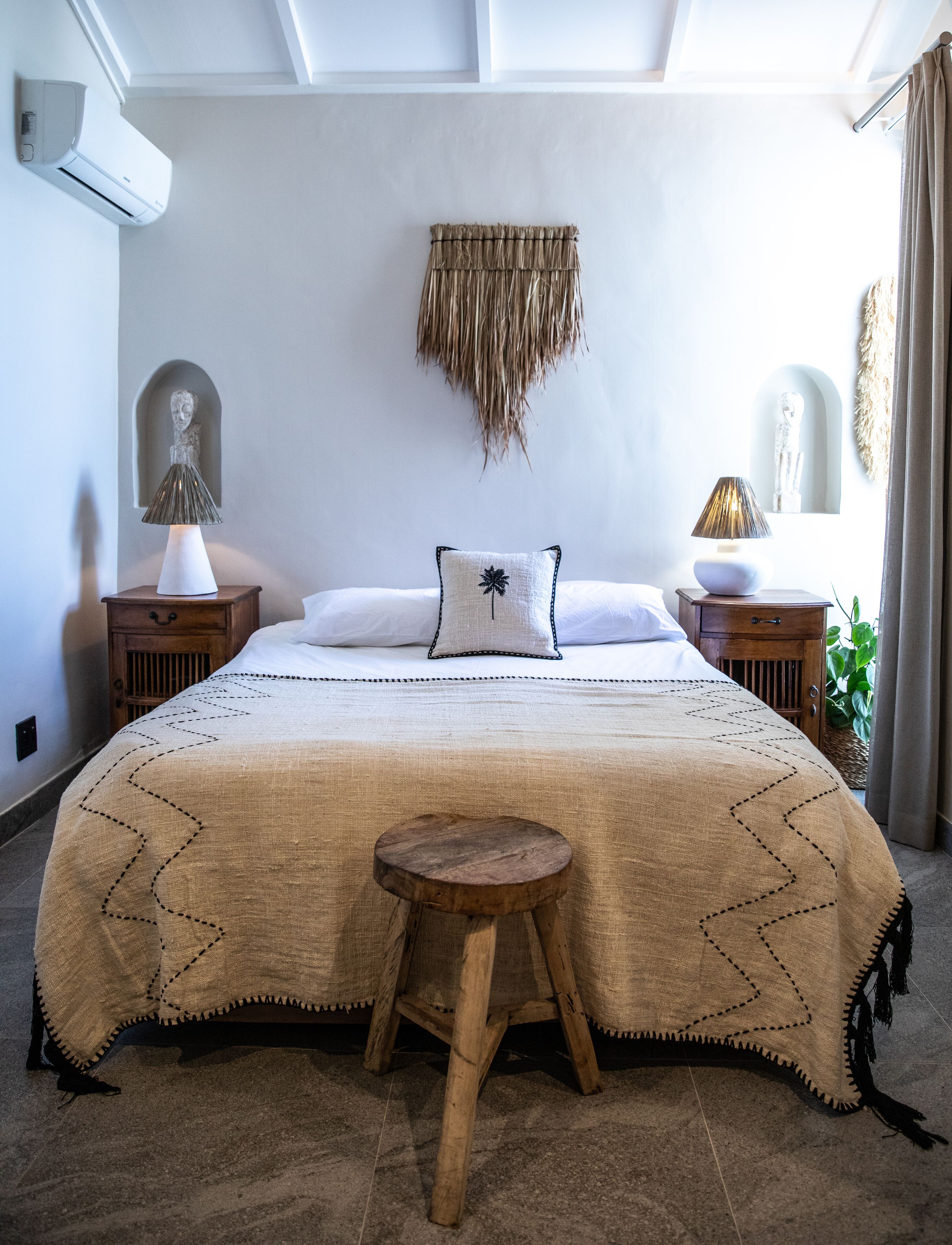Une chambre avec un grand lit, deux tables de chevet, un petit tabouret, un climatiseur fixé au mur et une décoration murale tissée.
