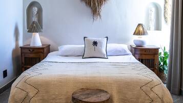 Une chambre avec un grand lit, deux tables de chevet, un petit tabouret, un climatiseur fixé au mur et une décoration murale tissée.