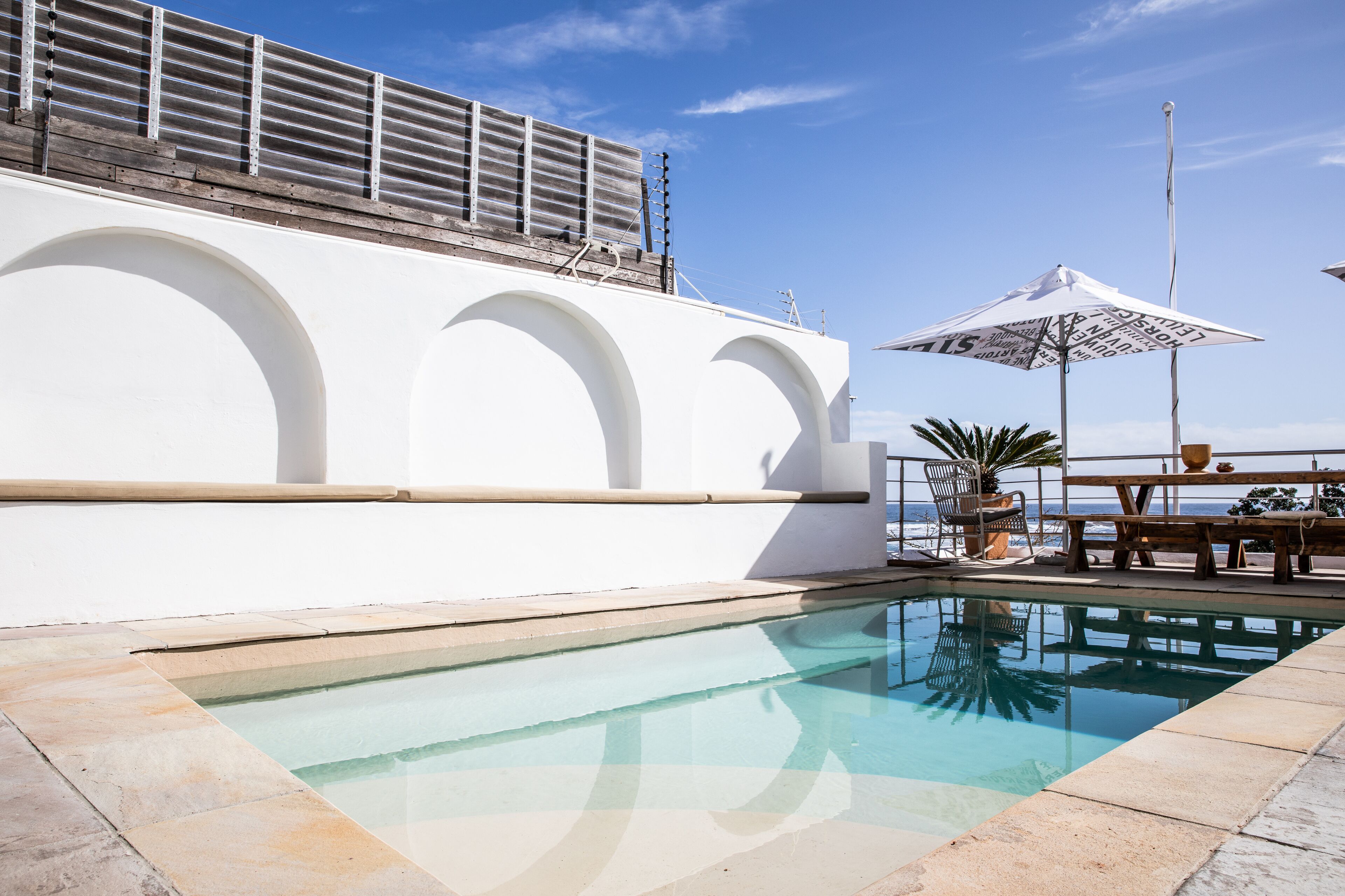 A swimming pool with a white wall and arched design, a wooden deck, and a table with chairs.