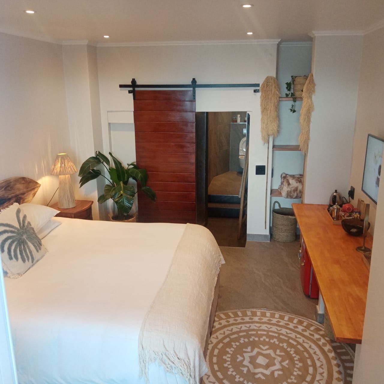 Een hotelkamer met een bed, nachtkastje, lamp, potplant en een schuifdeur naar een badkamer.