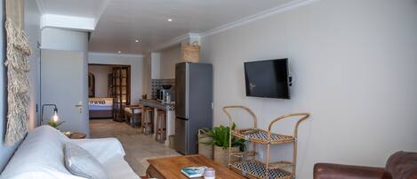 A living room with a sofa, coffee table, and a TV mounted on the wall. There is a kitchen area in the background with a refrigerator and a dining table.