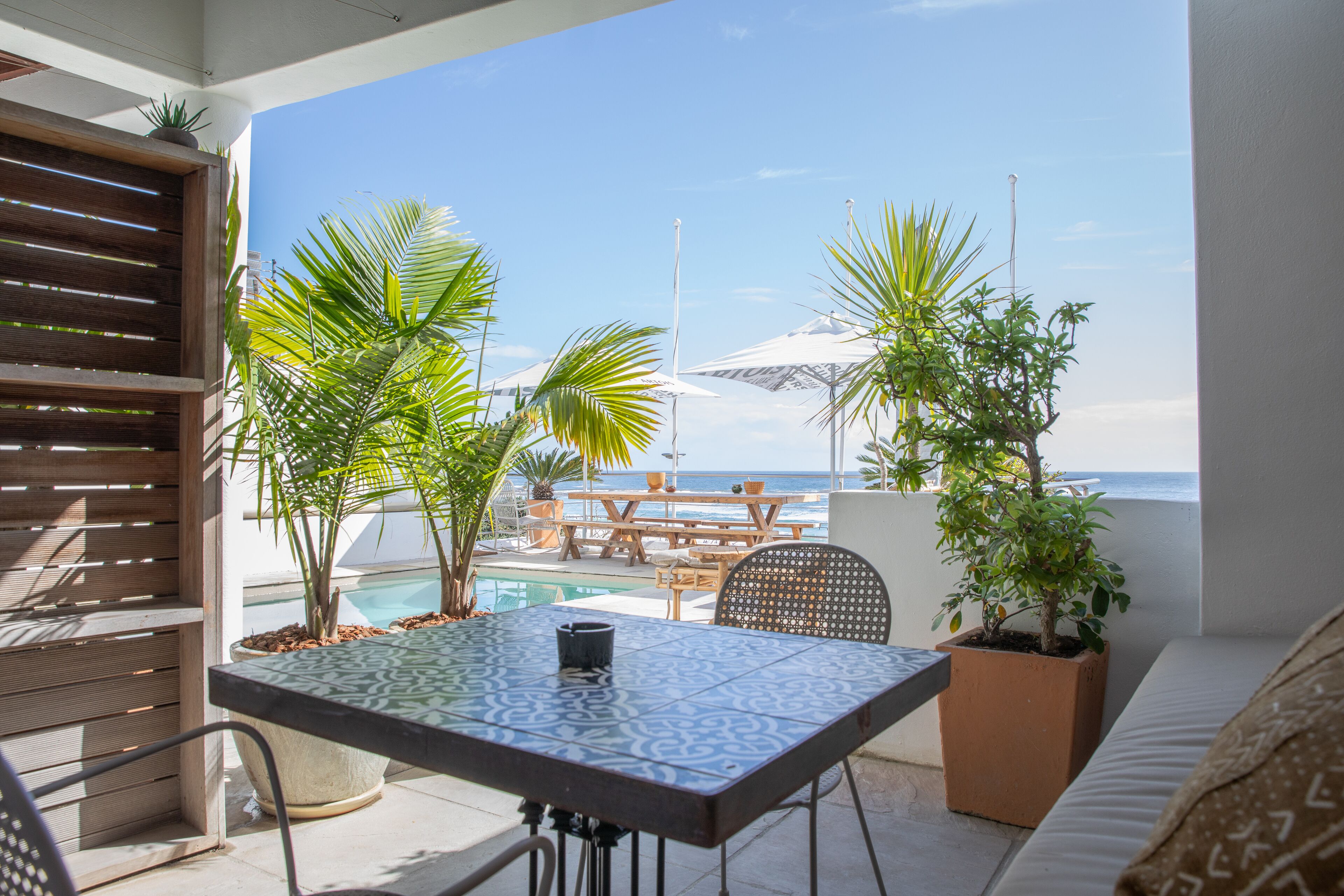 A patio with a table and chairs, surrounded by potted plants and overlooking a pool and ocean view.