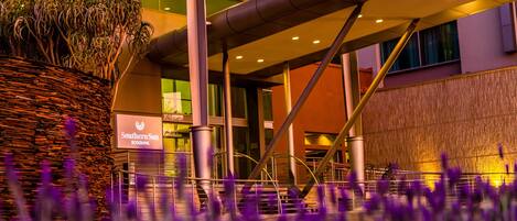 A modern building with a sign for Southern Sun Rosebank, a staircase, and a large planter with plants.