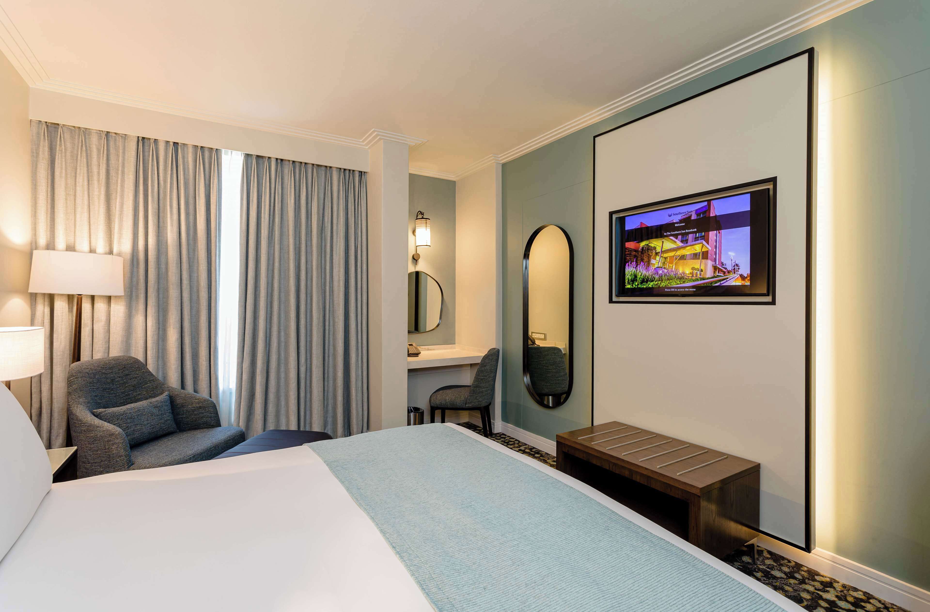 Una habitación de hotel con cama, una silla, un escritorio, un espejo y un televisor en la pared.