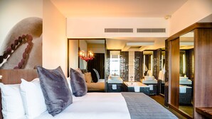 Habitación de hotel con una cama grande, un área de descanso con sofá y un baño con un espejo amplio y dos lavamanos.