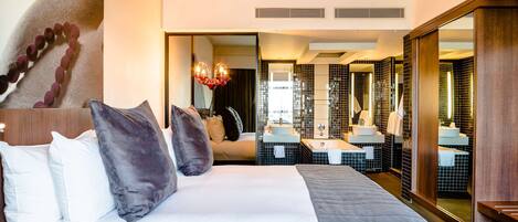Habitación de hotel con una cama grande, un área de descanso con sofá y un baño con un espejo amplio y dos lavamanos.