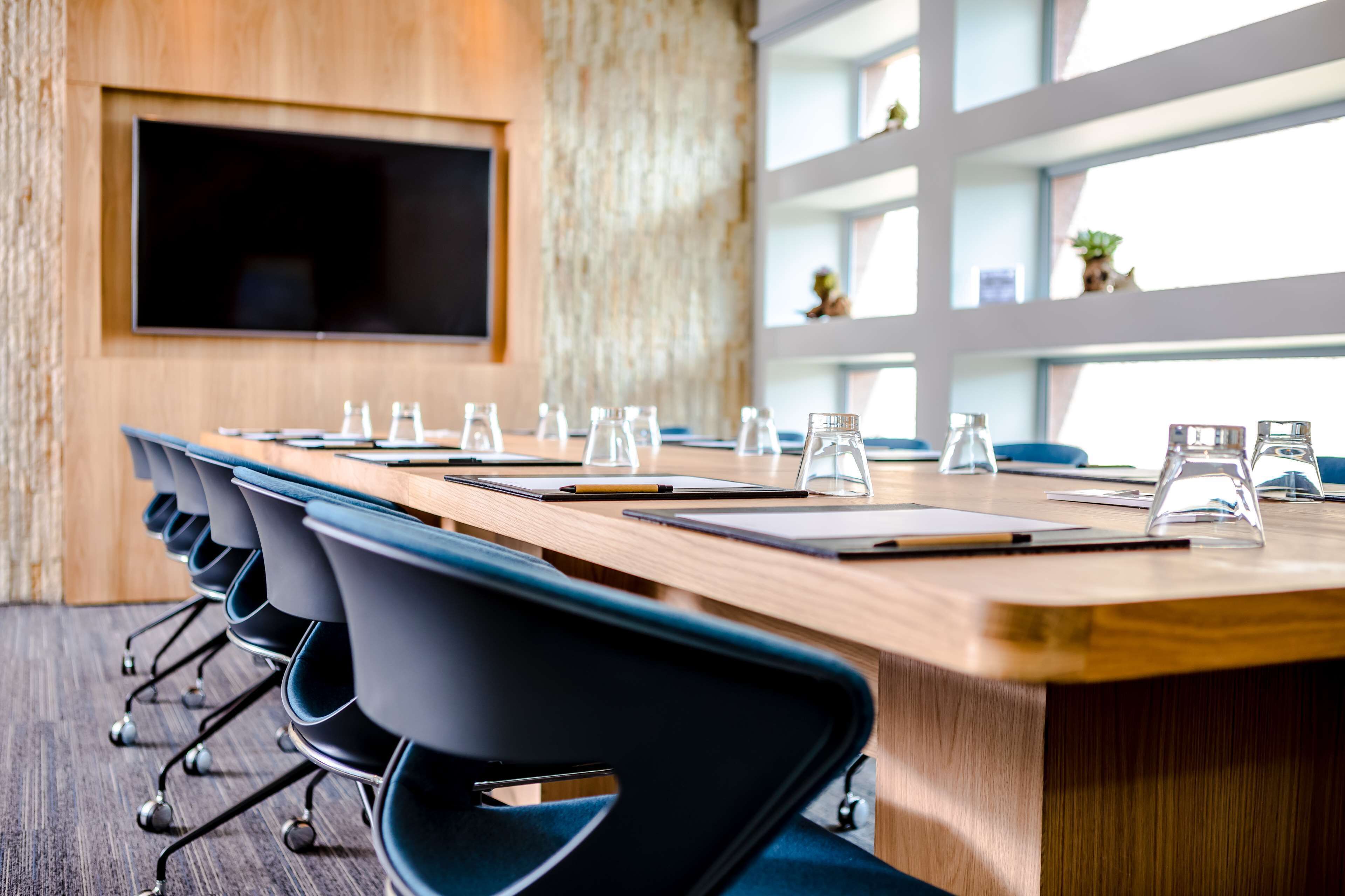 A conference room with a long table, chairs, a TV screen, and a window with natural light.