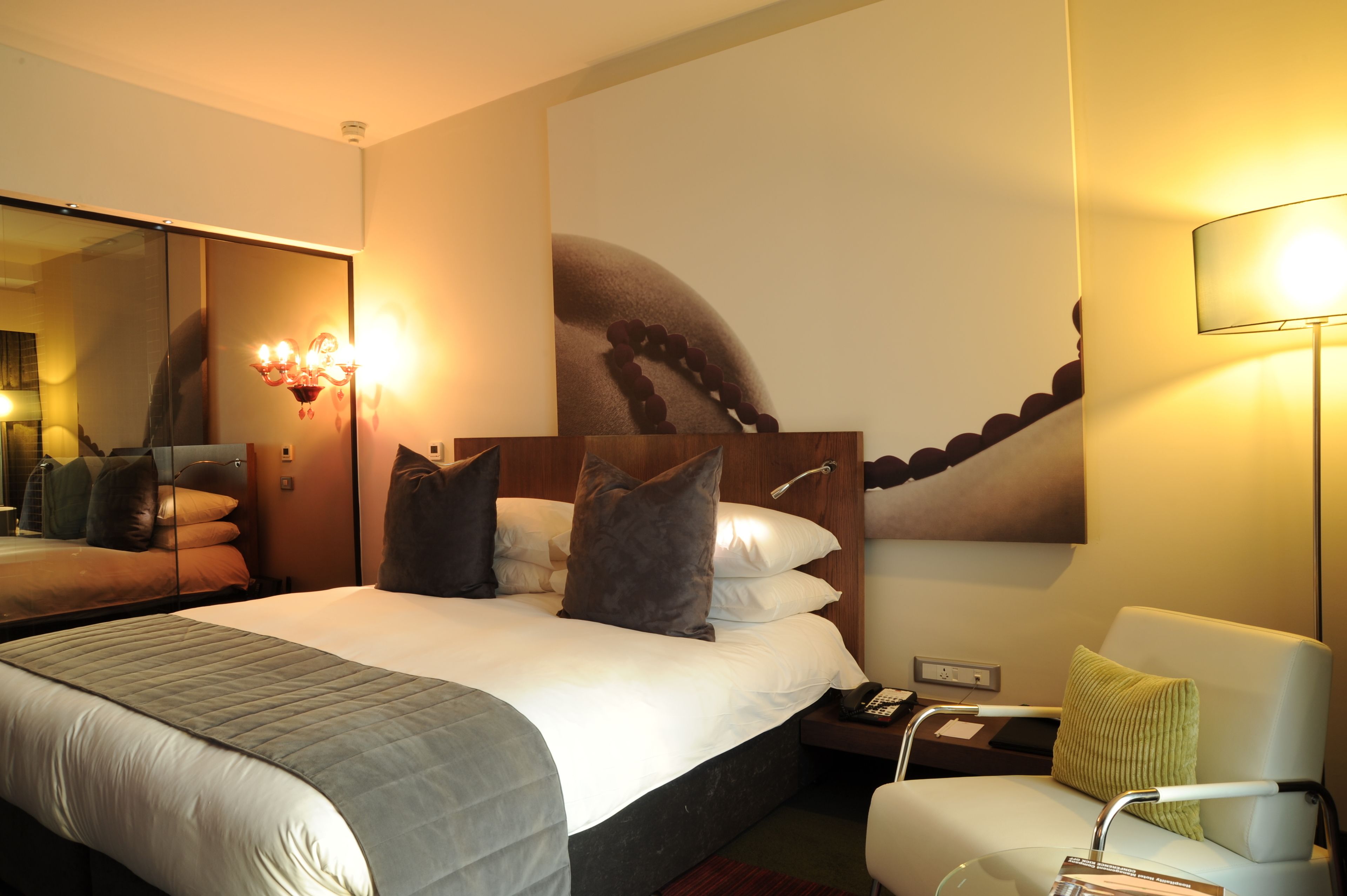 A hotel room with a large bed, a nightstand, a chair, a mirror, and a wall with a large artwork.