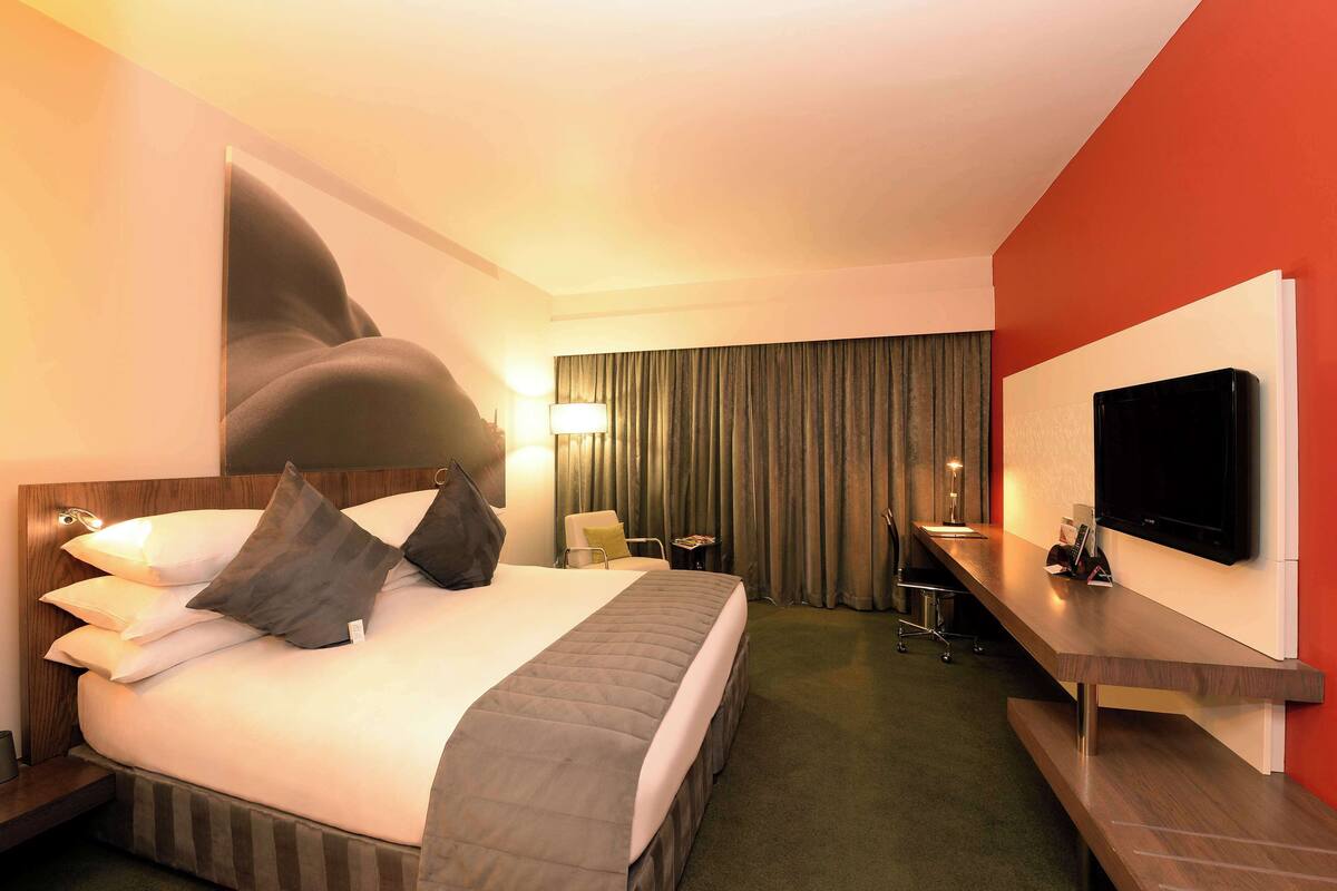 A hotel room with a large bed, a desk, a flat-screen TV, and a view of the city.