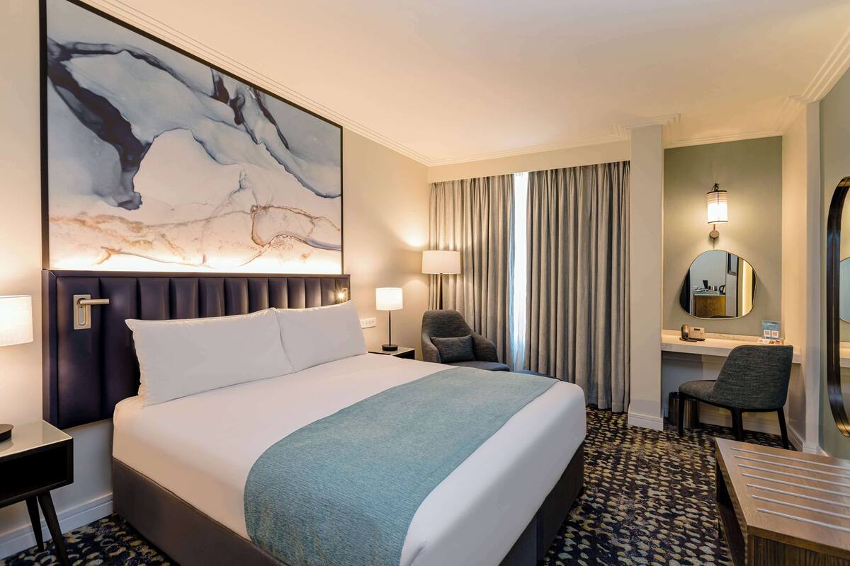 A hotel room with a large bed, a desk, a chair, a desk lamp, a mirror, and a window with curtains.