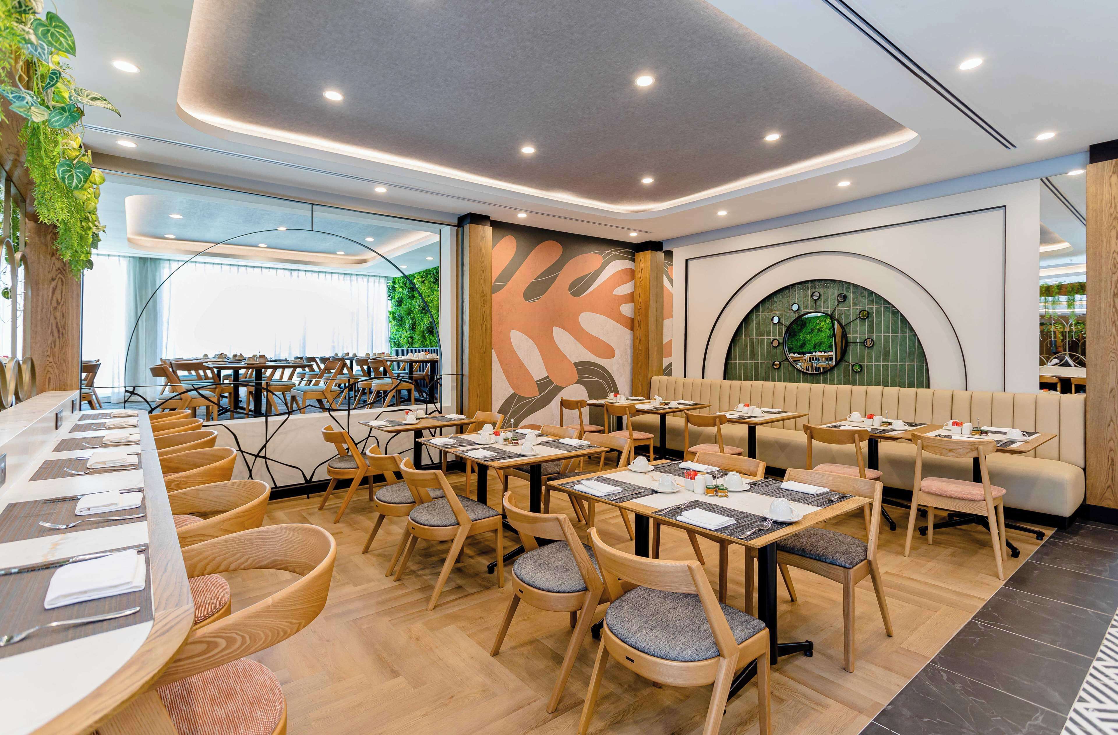A modern restaurant with wooden tables and chairs, a large mural, and a circular green wall feature.