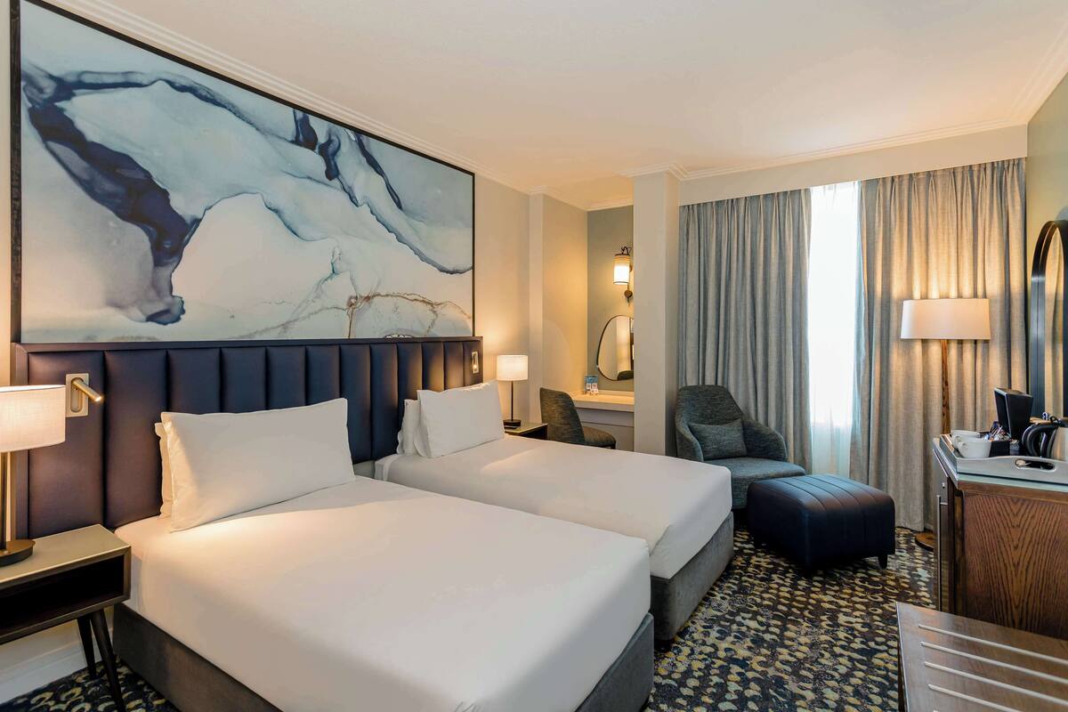 Habitación de hotel con dos camas, un cuadro abstracto grande, un escritorio con lámpara y vistas al exterior a través de cortinas.