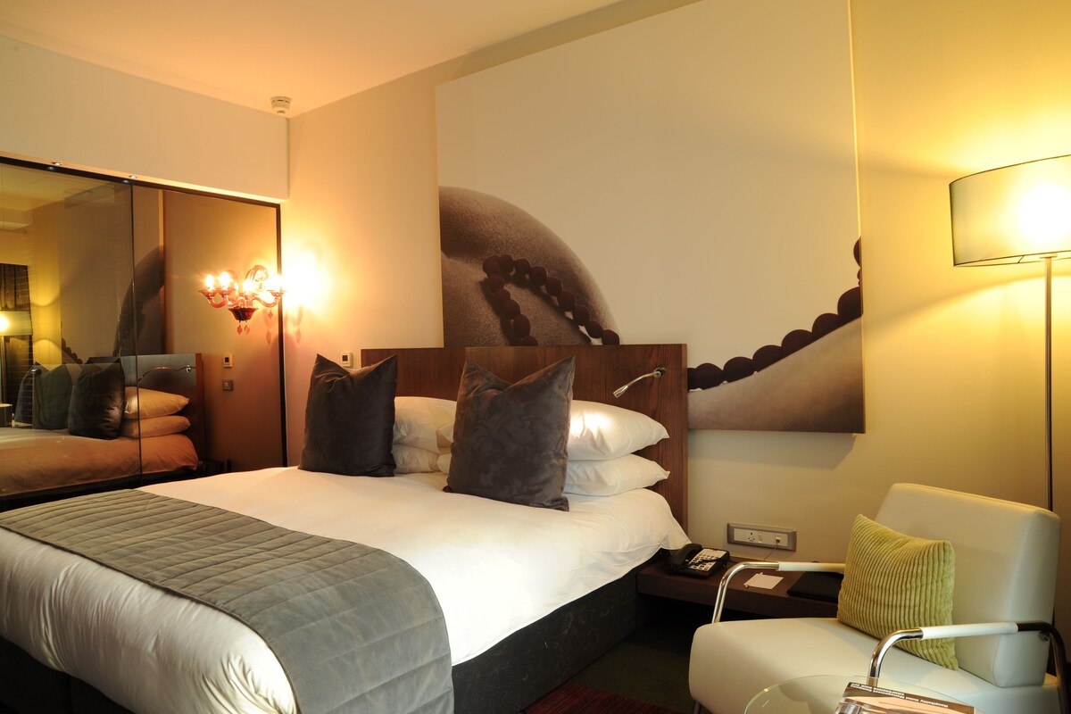 A hotel room with a large bed, a bedside table with a lamp, a chair, and a wall with a large artwork.