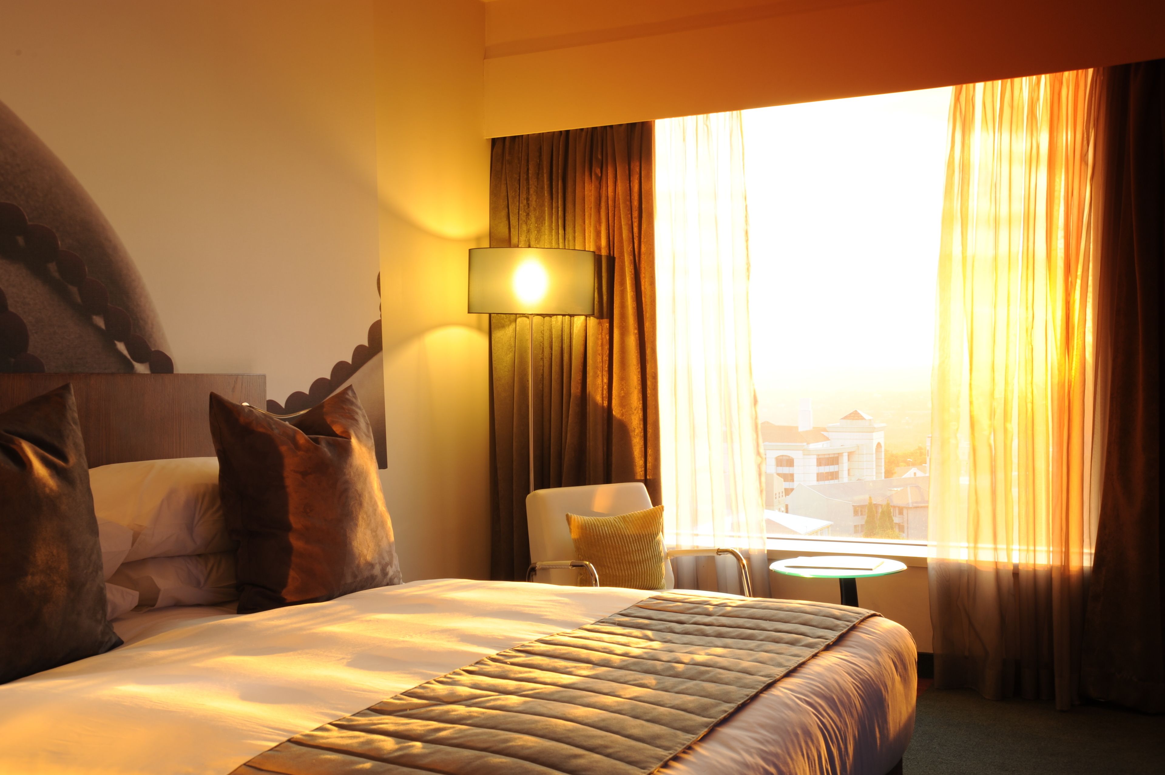 A hotel room with a bed, pillows, a lamp, a chair, a small table, and a window with curtains.