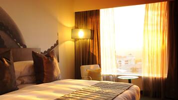 Ein Hotelzimmer mit Bett, Kissen, Lampe, Stuhl, einem kleinen Tisch und einem Fenster mit Vorhängen.