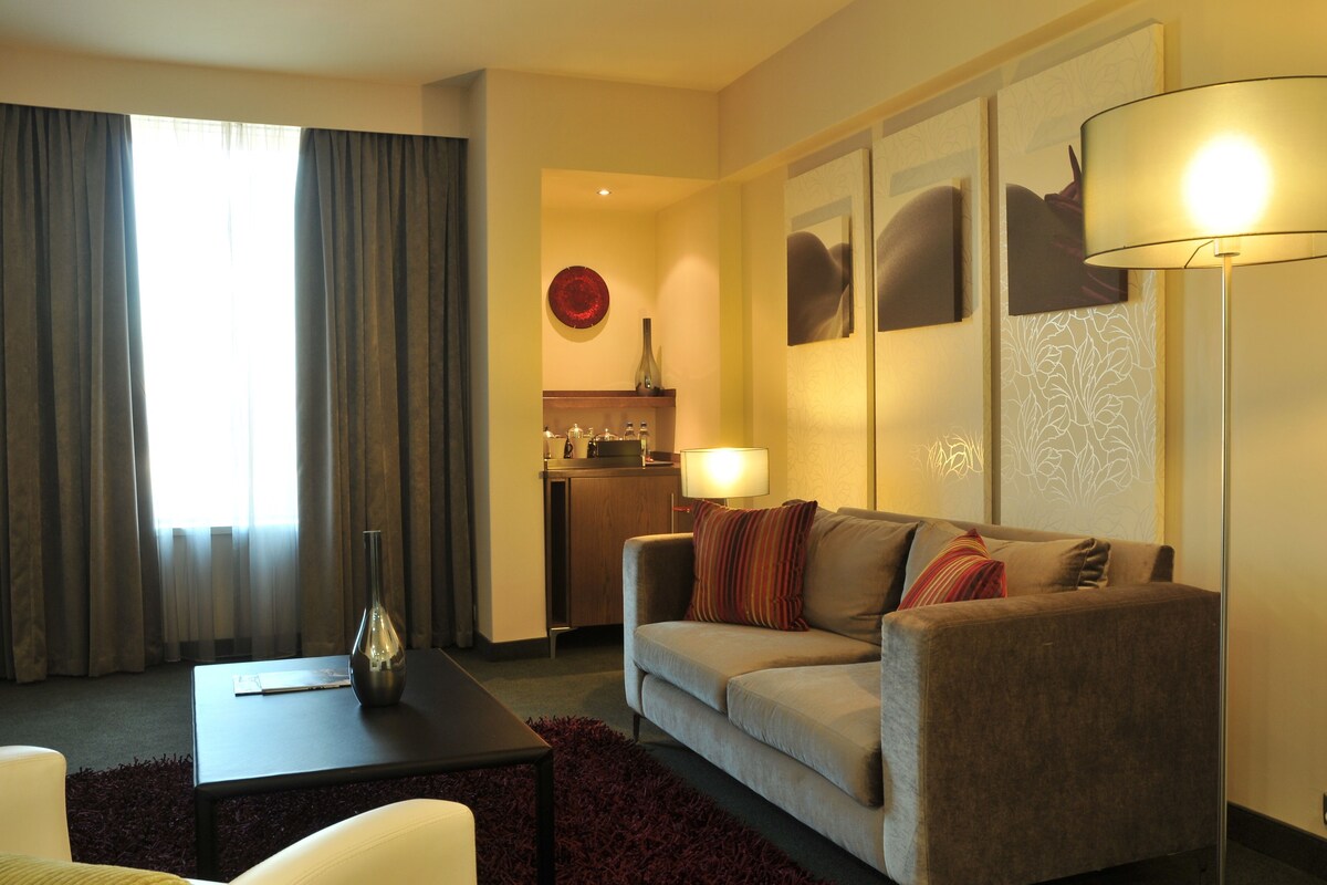 A hotel room with a sofa, a small table, a lamp, a vase, and a view of the kitchen.