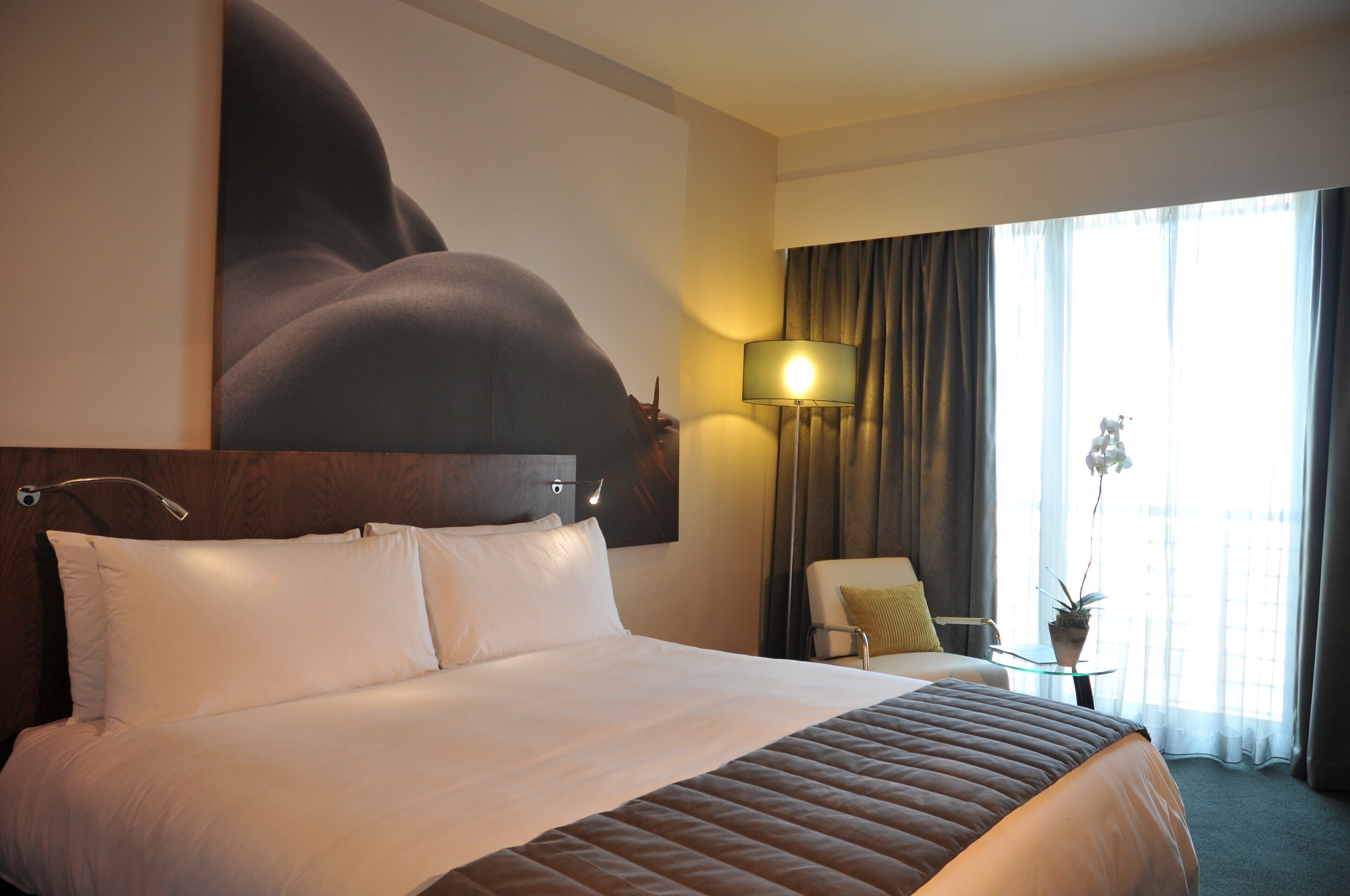 A hotel room with a large bed, a desk with a lamp, a chair, and a window with curtains.