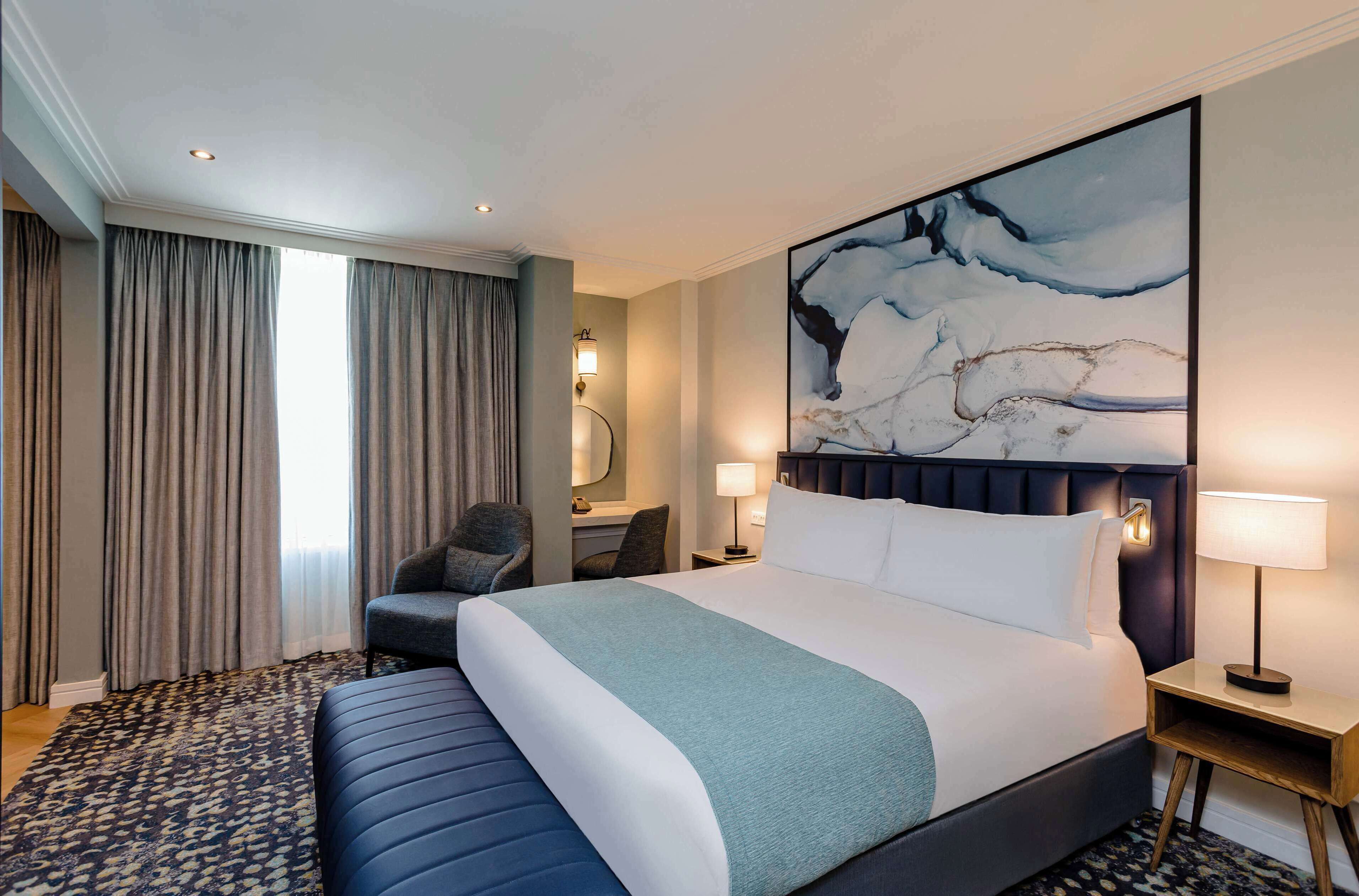 A hotel room with a large bed, a desk with a chair, a desk lamp, and a large abstract painting on the wall.