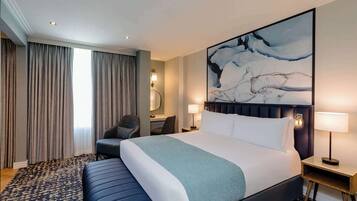 A hotel room with a large bed, a desk with a chair, a desk lamp, and a large abstract painting on the wall.
