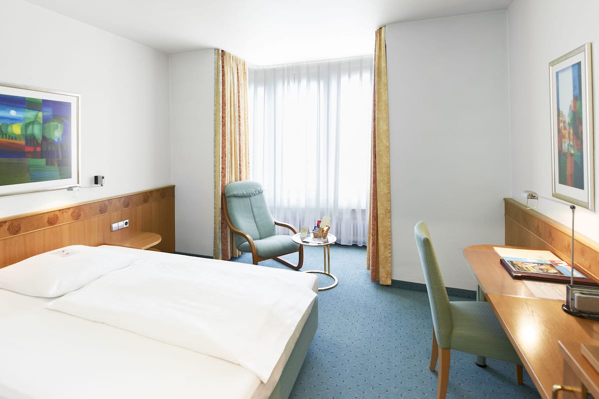 Habitación de hotel con cama, escritorio, silla, televisor y ventana con cortinas.