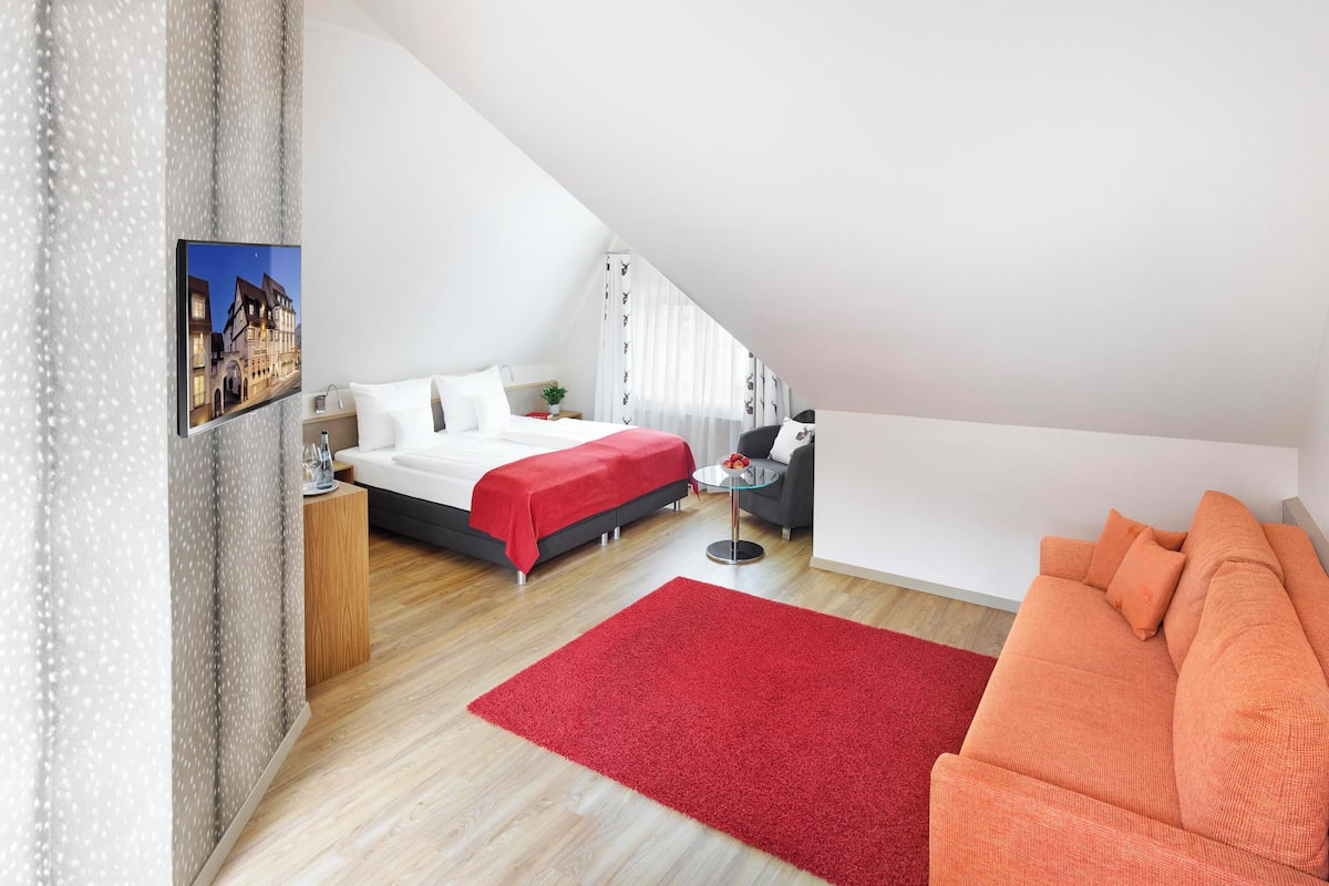 Un dormitorio moderno con cama, sofá, alfombra roja y un cuadro en la pared.
