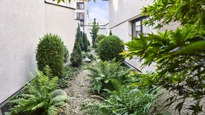 Un estrecho callejón con un sendero de grava, flanqueado por densa vegetación y árboles altos, que conduce a un edificio con fachada blanca y varias ventanas.