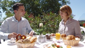 Una pareja desayunando al aire libre, con una mesa dispuesta que incluye pan, huevos y fruta.