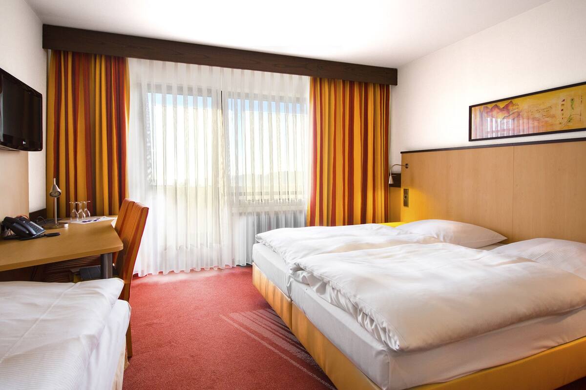 A hotel room with two beds, a desk with a telephone and glasses, a chair, and a TV on the wall.