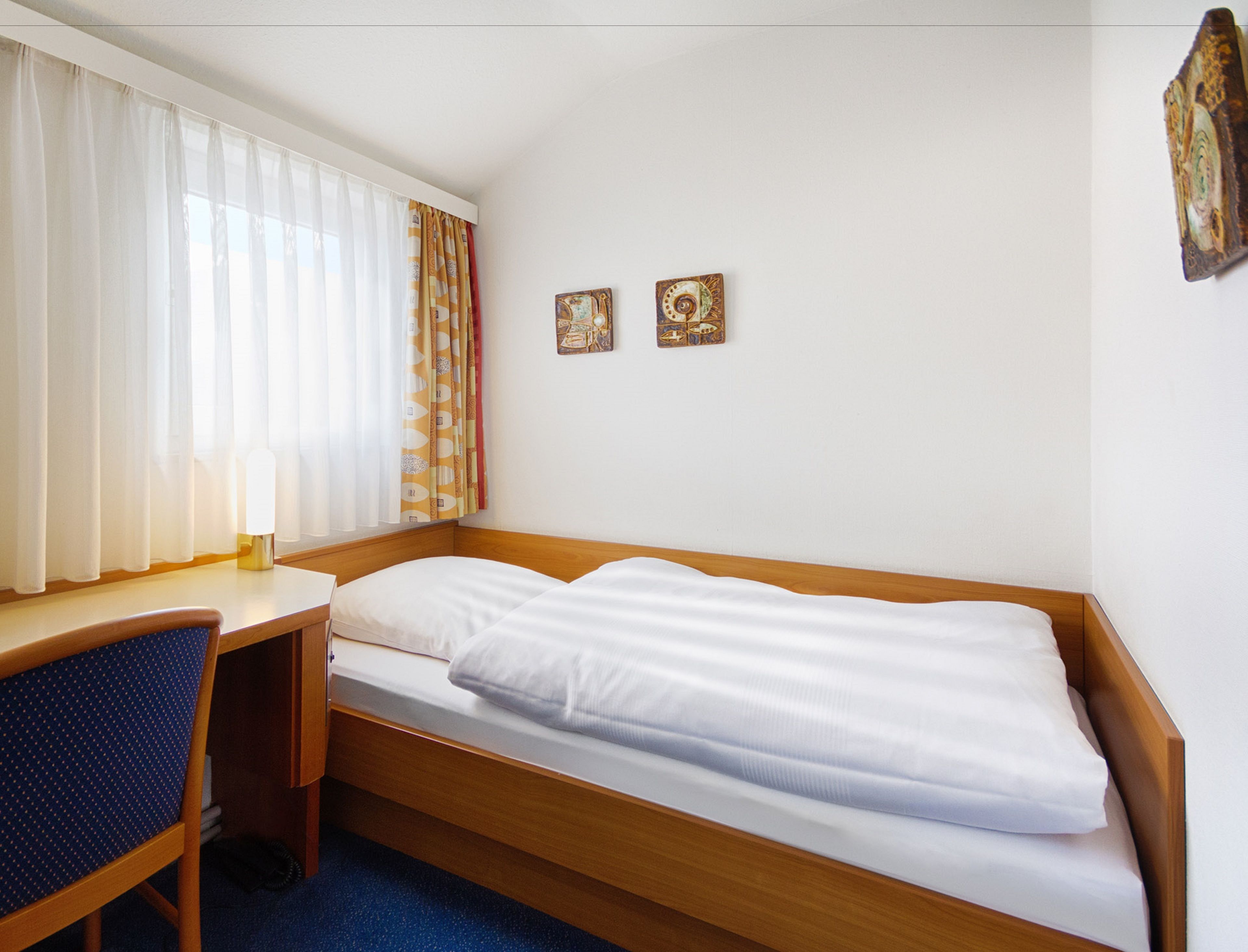 Una habitación pequeña con cama individual, cabecera de madera, un escritorio con silla y dos cuadros en la pared.