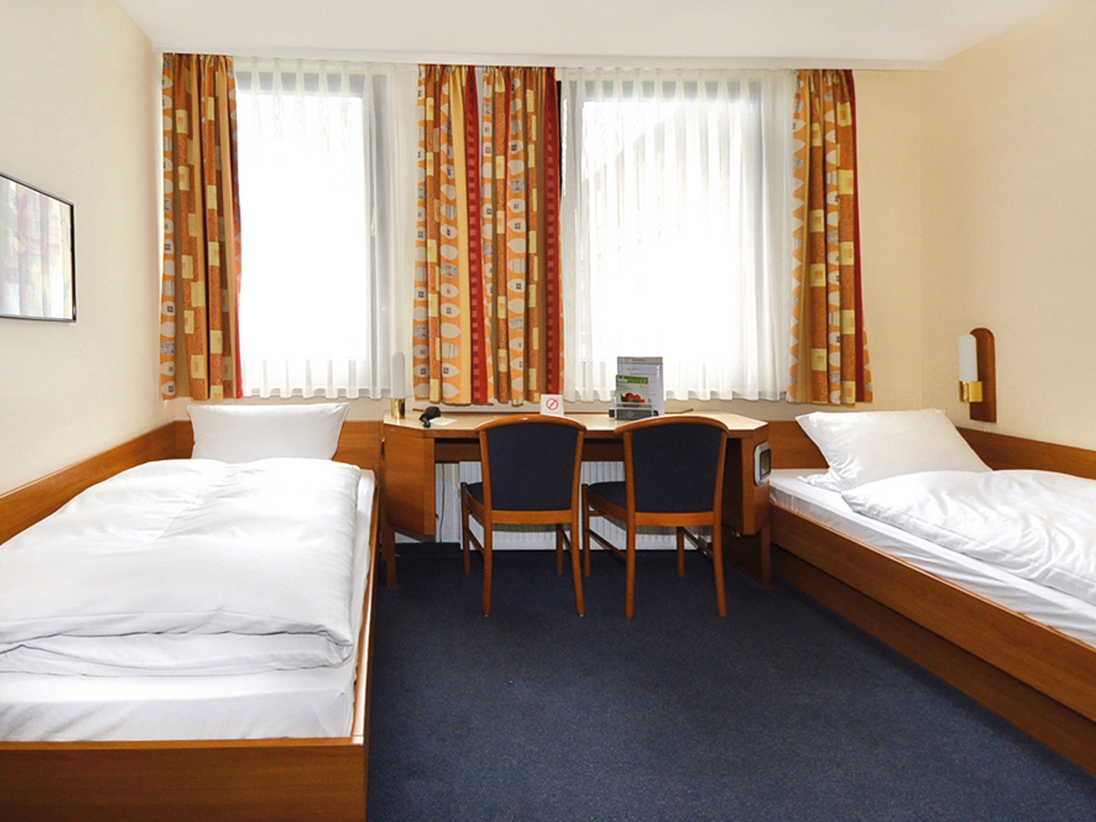 A hotel room with two single beds, a desk with a chair, and a window with curtains.