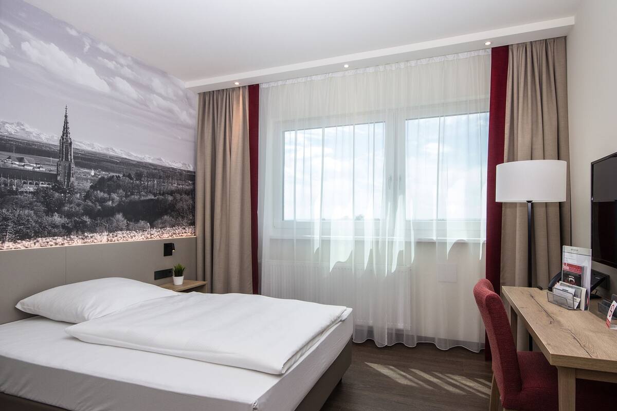 A hotel room with a bed, a desk, a chair, a lamp, and a large window with curtains.