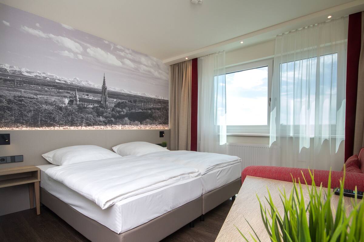 A modern hotel room with a large bed, a small bedside table, a red sofa, and a large window with curtains.