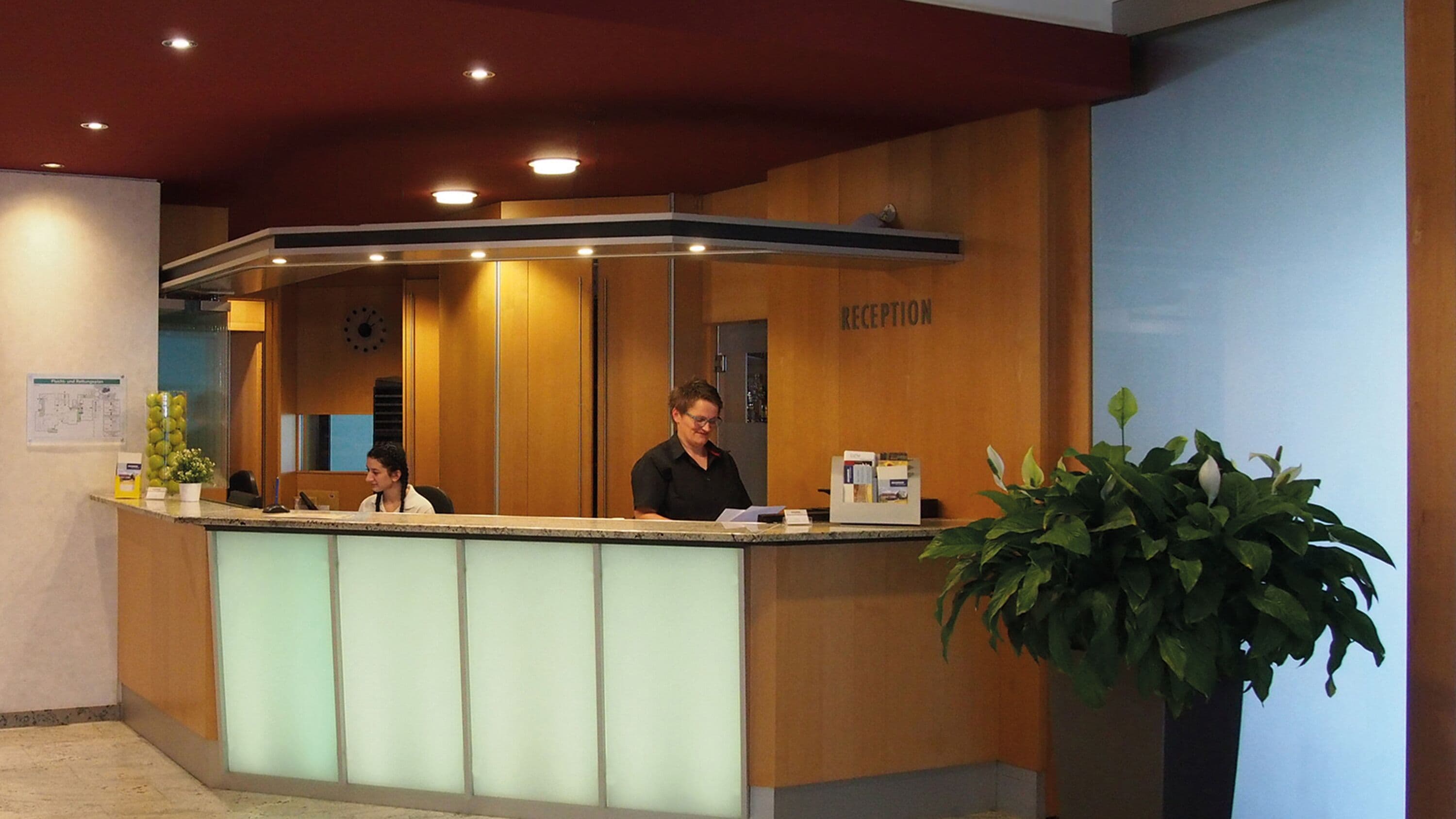 A reception desk with two staff members, a clock, and a reception sign.