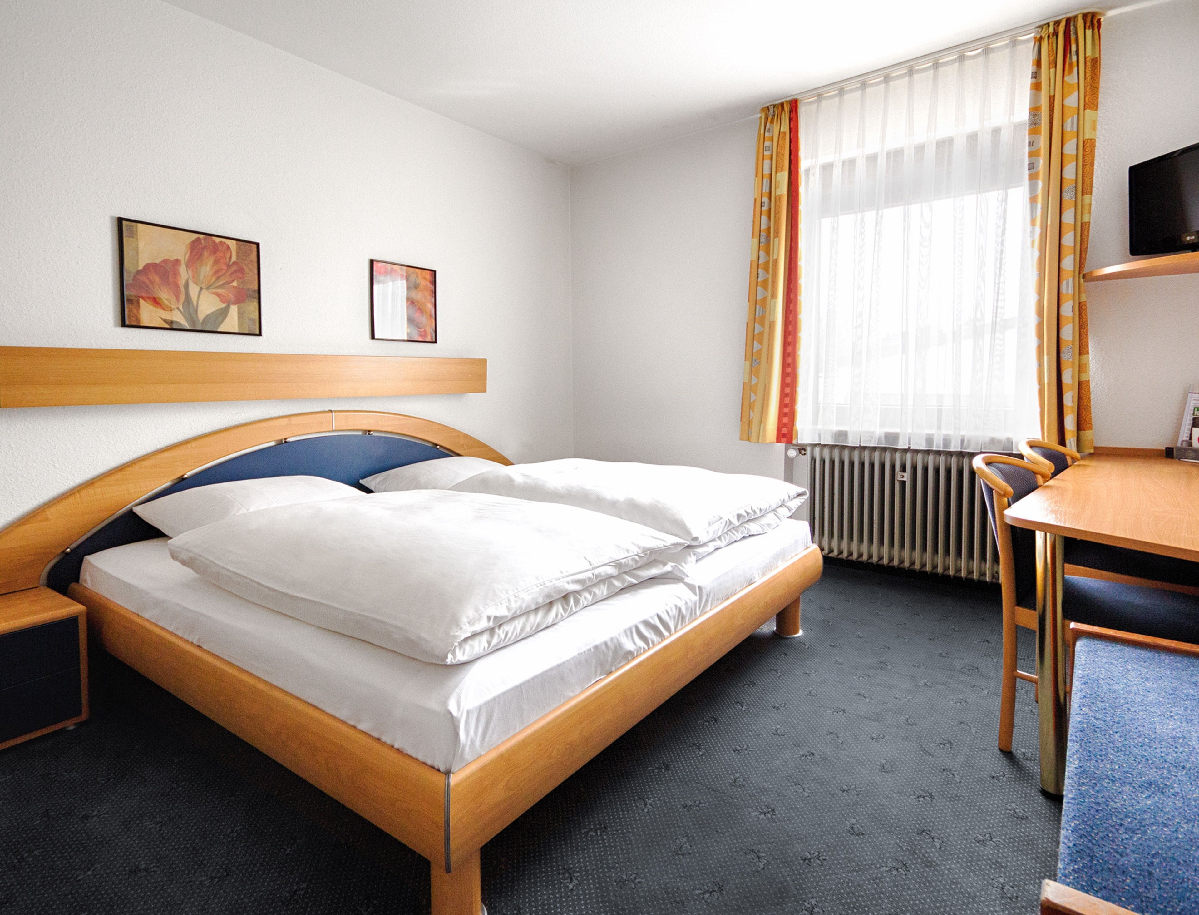 A hotel room with a wooden bed, a desk with a chair, a radiator, and two framed pictures on the wall.