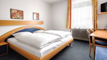 A hotel room with a wooden bed, a desk with a chair, a radiator, and two framed pictures on the wall.