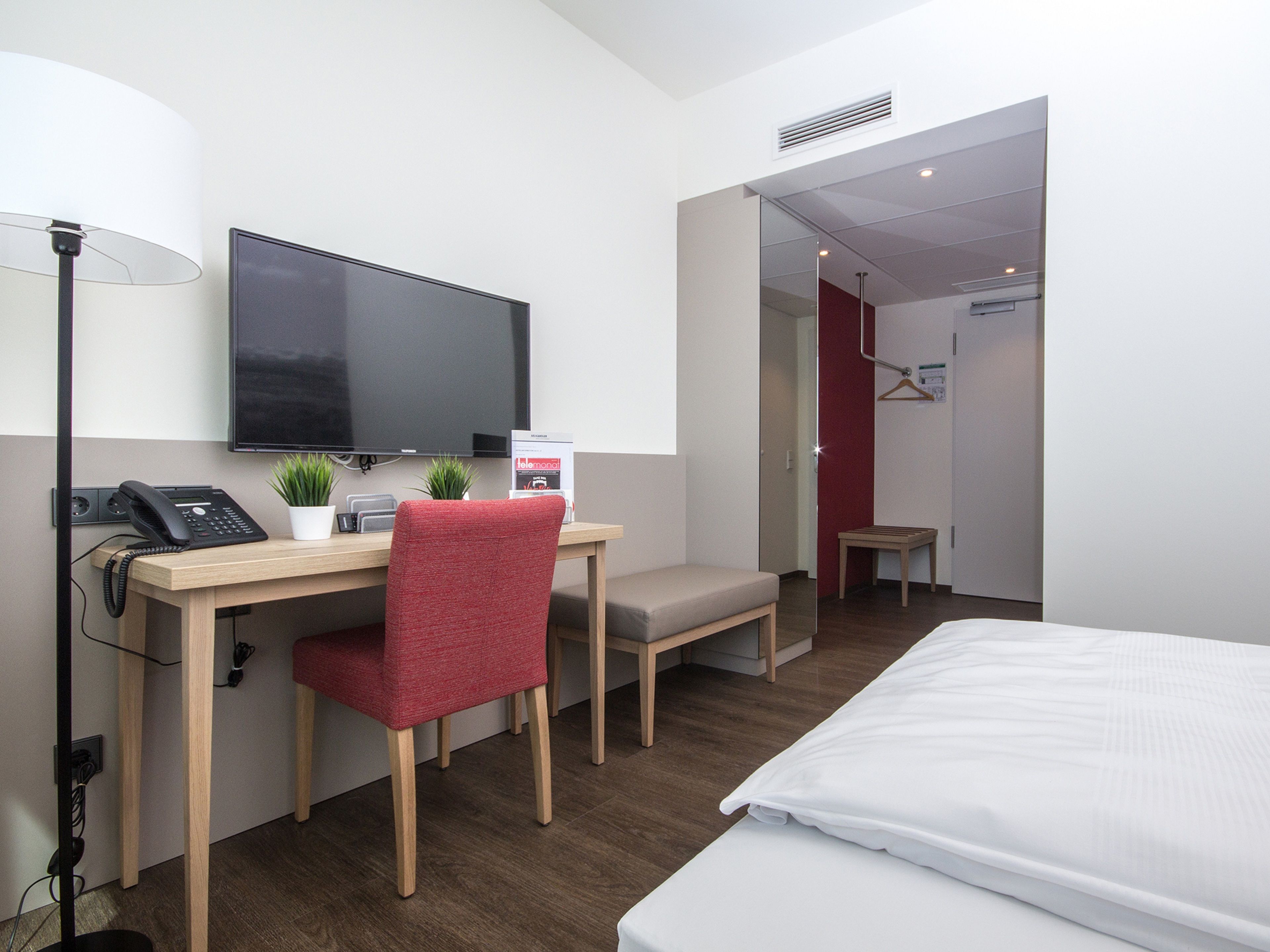 Habitación de hotel con cama, escritorio con silla, televisión, teléfono y una planta pequeña.