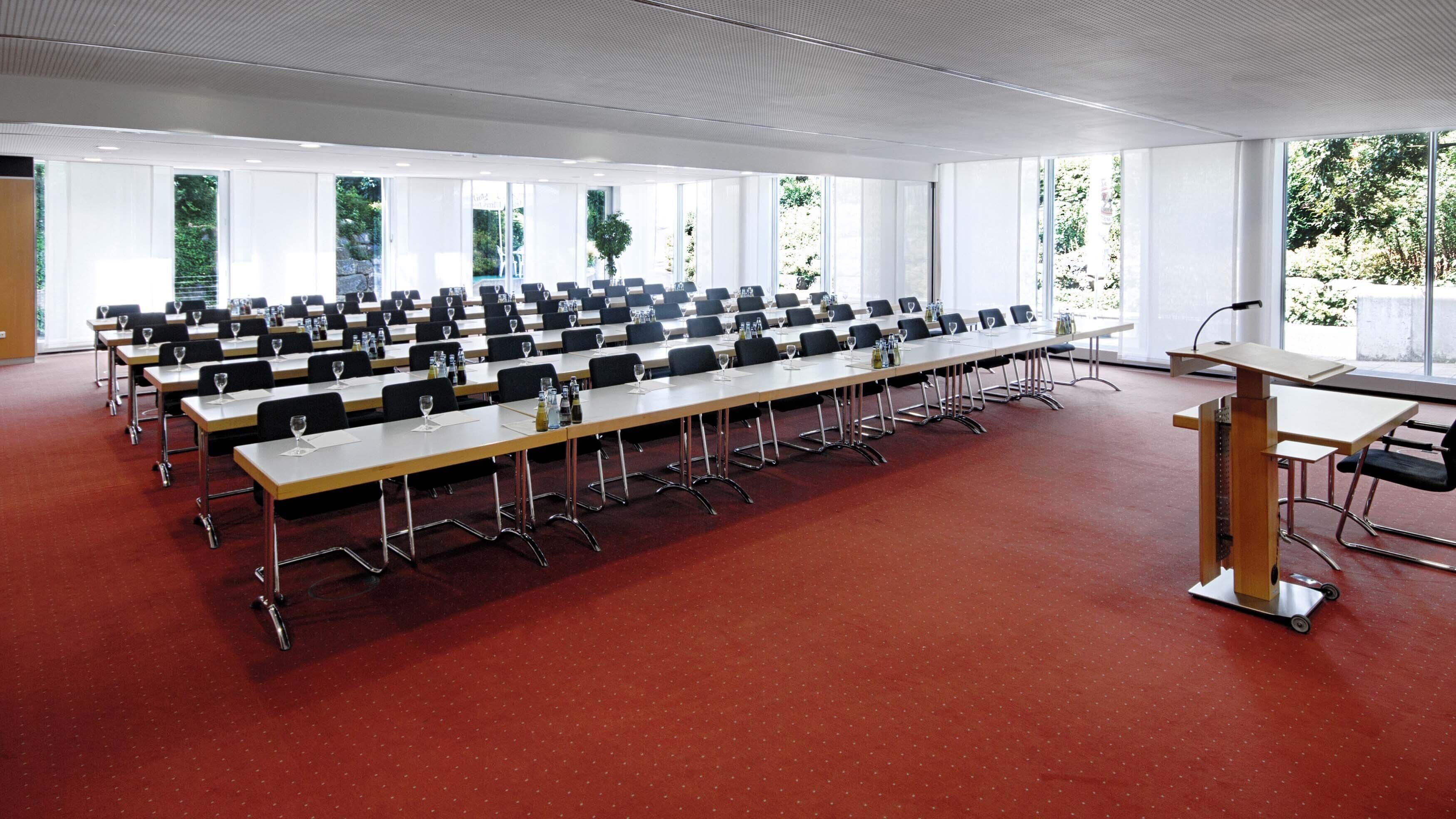 Un salón de conferencias con filas de mesas y sillas, ventanas amplias y un atril.