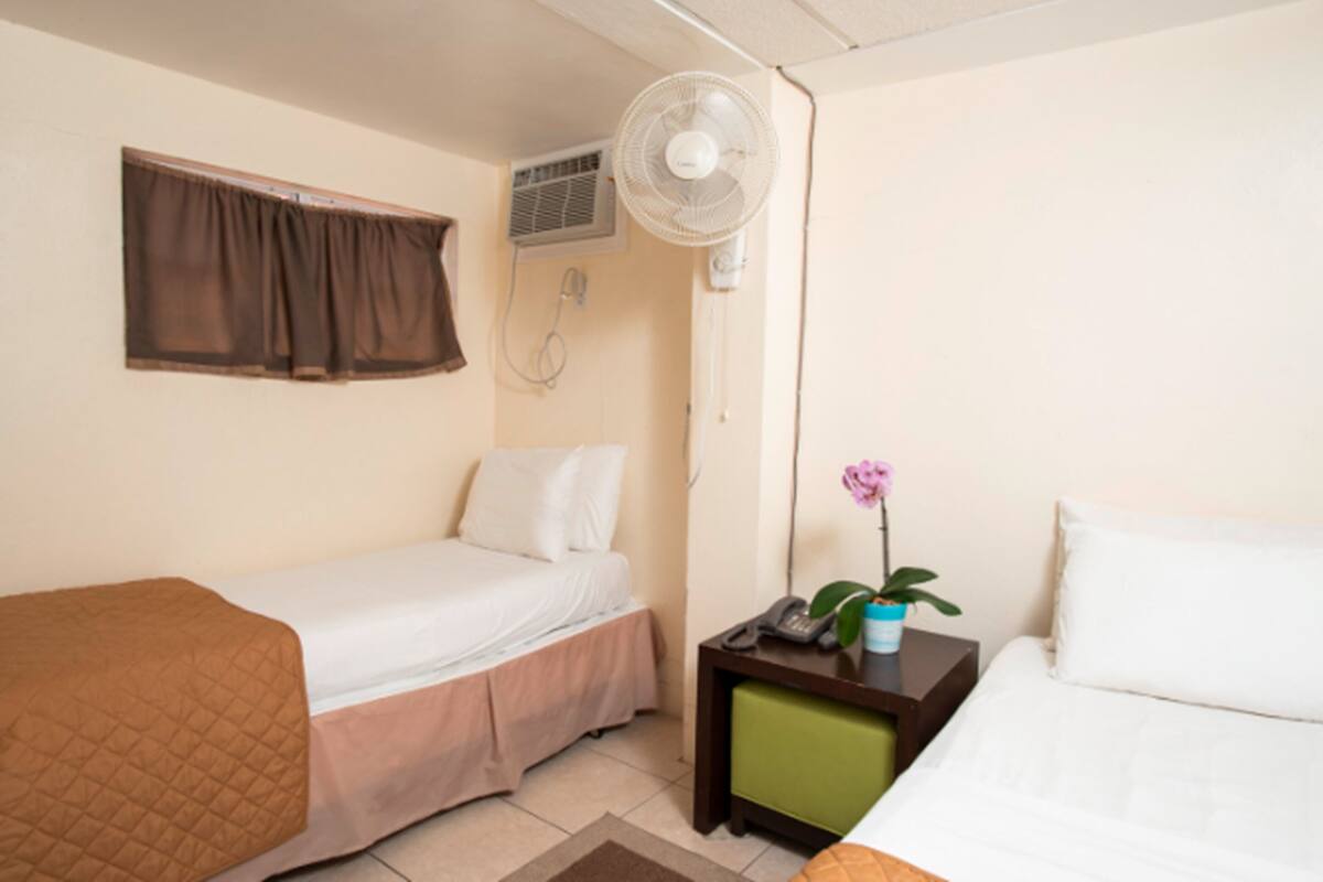 A small, clean room with two beds, a nightstand, a fan, and a window with curtains.