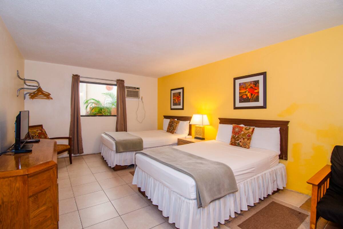 A hotel room with two beds, a desk, a chair, a window with curtains, and a wall-mounted air conditioner.
