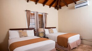 A hotel room with two beds, a nightstand, a lamp, and an air conditioning unit. - Bunker Hill Hotel (St. Thomas)