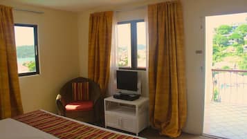 A hotel room with a bed, a television on a small stand, a chair, and a balcony with a view of a hillside.