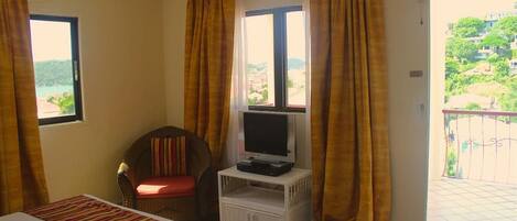 A hotel room with a bed, a television on a small stand, a chair, and a balcony with a view of a hillside.