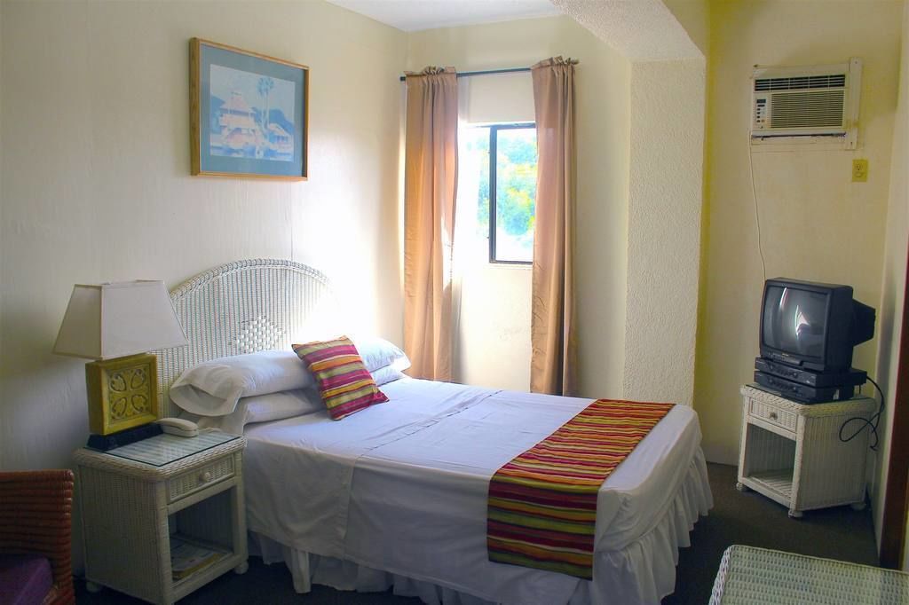 Quarto de hotel com cama, televisão, mesinha, mesa de cabeceira e janela com cortinas.