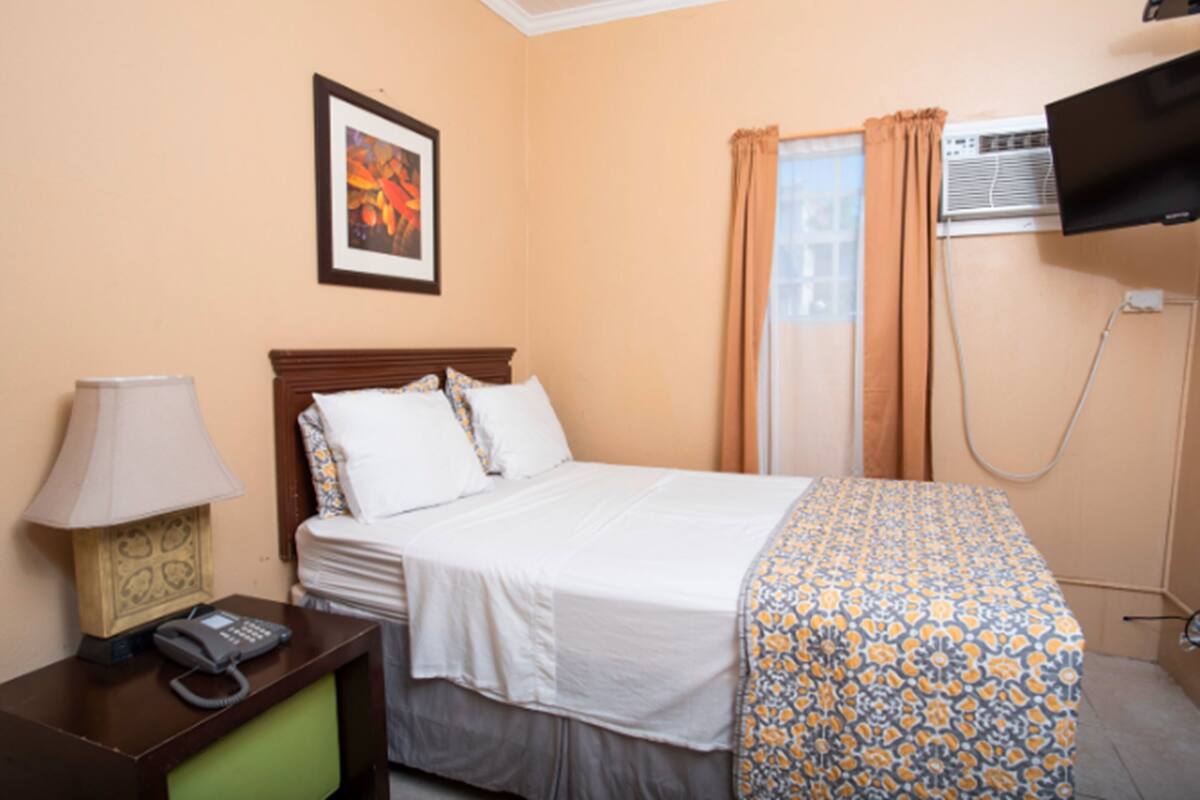 A hotel room with a bed, a bedside table with a lamp and phone, a framed picture on the wall, and an air conditioning unit.