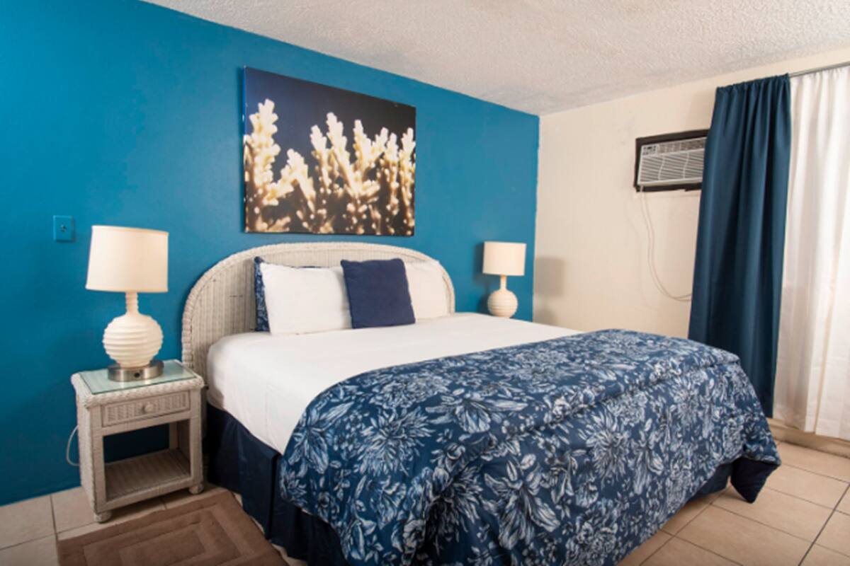 A hotel room with a bed, two bedside lamps, a wall-mounted air conditioner, and a window with curtains.